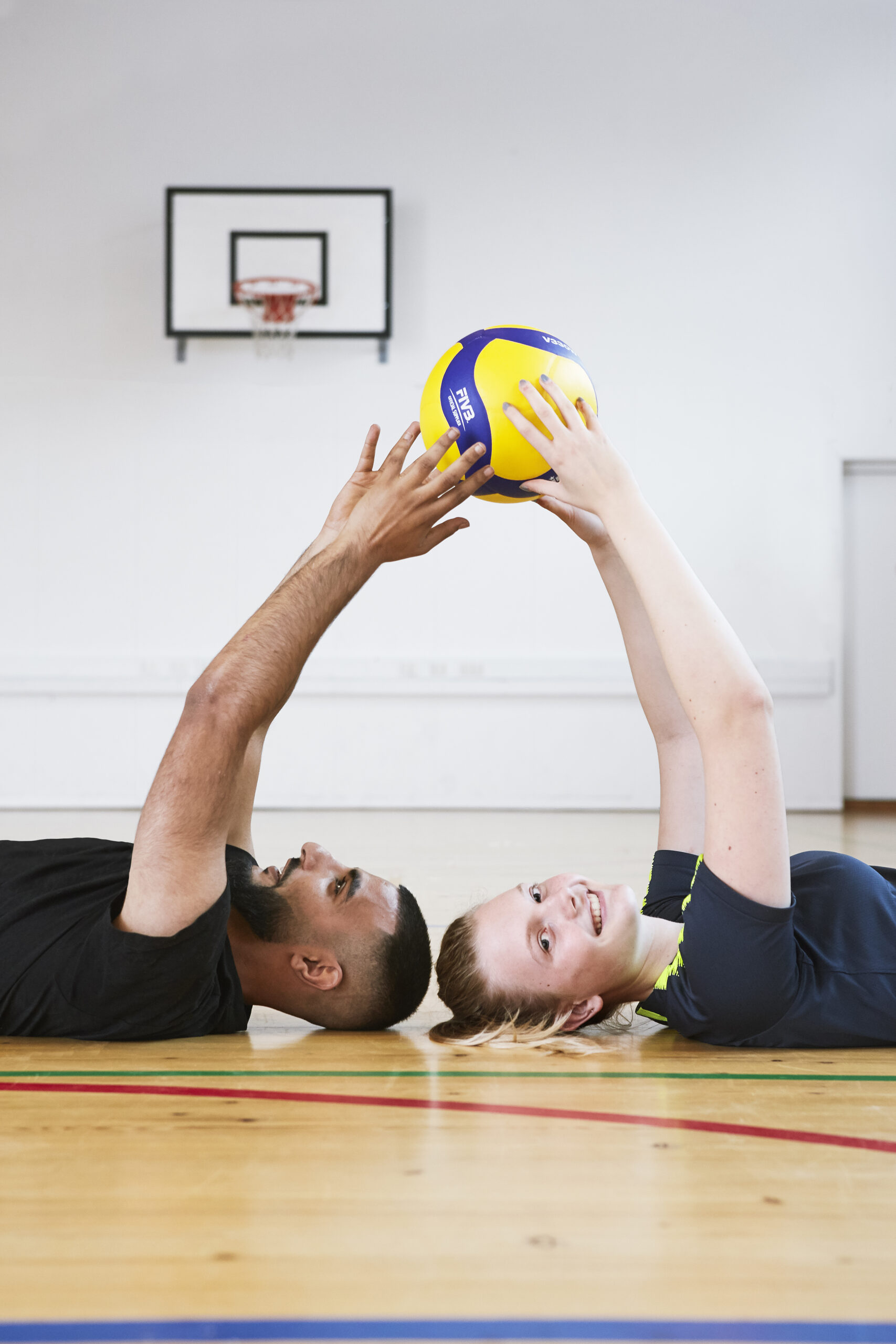 To elever ligger på et gulv i en gymnastiksal og griber ud efter en bold